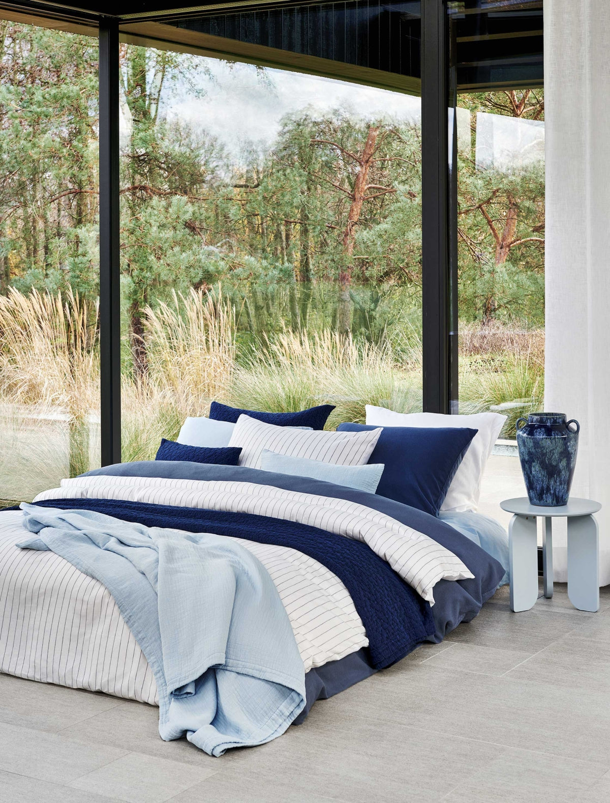 Een bed met een donkerblauw dekbedovertrek, een wit dekbedovertrek met dunne blauwe strepen en op het bed is een lichtblauw plaid gedrapeerd